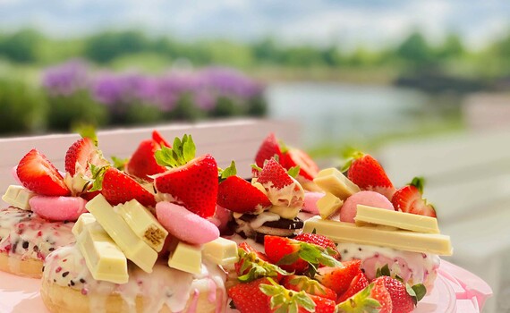 Bunt verzierte Donuts stehen auf dem Bistrotisch des Restaurants Zauberküche auf der Terrasse des Jahrtausendturms vor der Kulisse des Angersees.