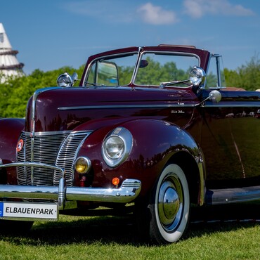 Oldtimer steht auf der Wiese im Elbauenpark Magdeburg. Im Hintergrund sieht man von Bäumen verdeckt den Jahrtausendturm.