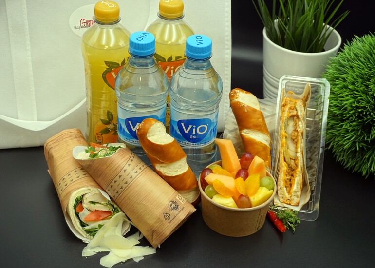 Weiße Picknicktasche mit 2 Flaschen gelber Limonade, 2 Flaschen Wasser, Sandwiches, Wraps und einem Obstsalat.